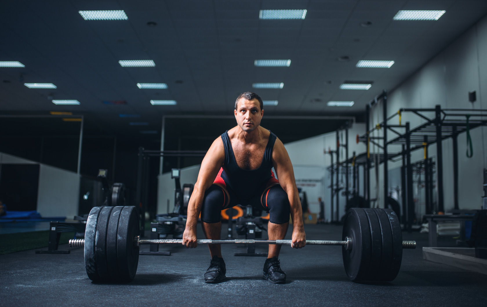 Man in a gym crouched and gripping a loaded barbell before a deadlift, wearing a tank top and knee sleeves