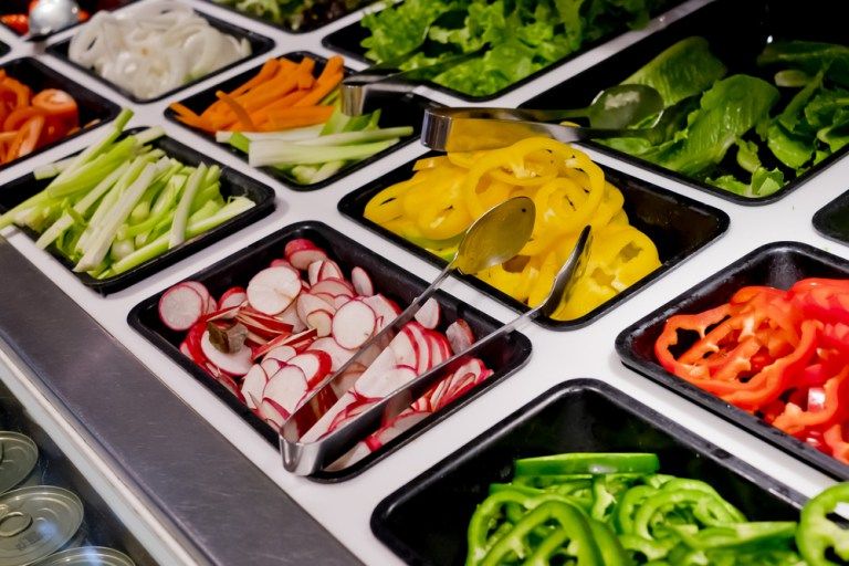 High-angle view of a salad bar with black trays of sliced radishes, bell peppers, carrots, onions, and lettuce with serving tongs