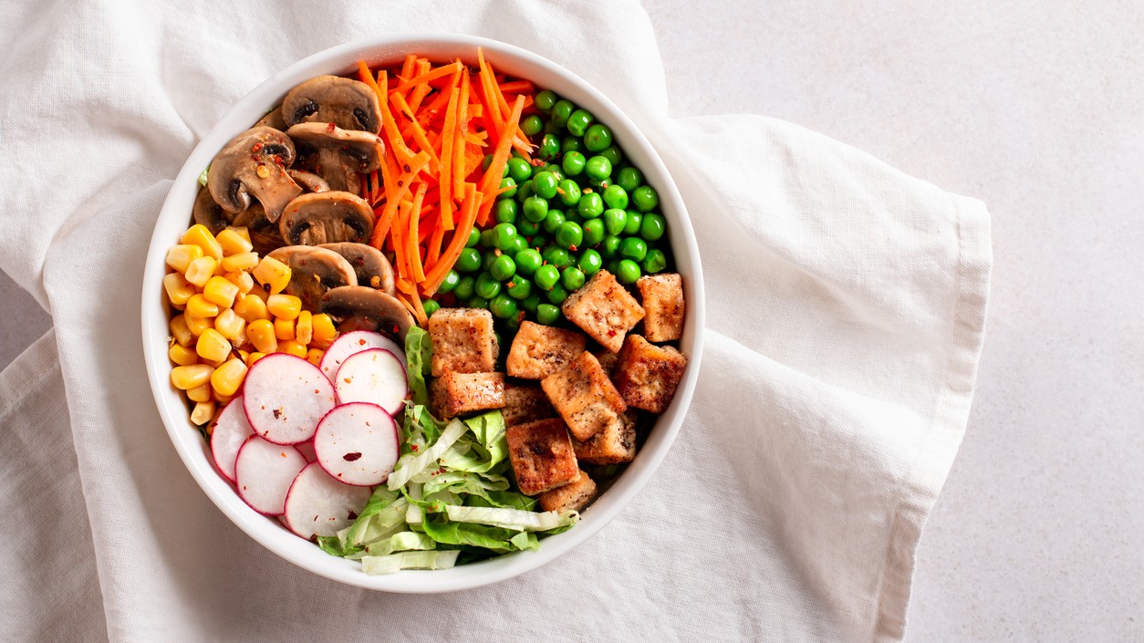 Top-down view of a white bowl with pan-seared tofu, mushrooms, carrots, peas, corn, radishes, and shredded lettuce